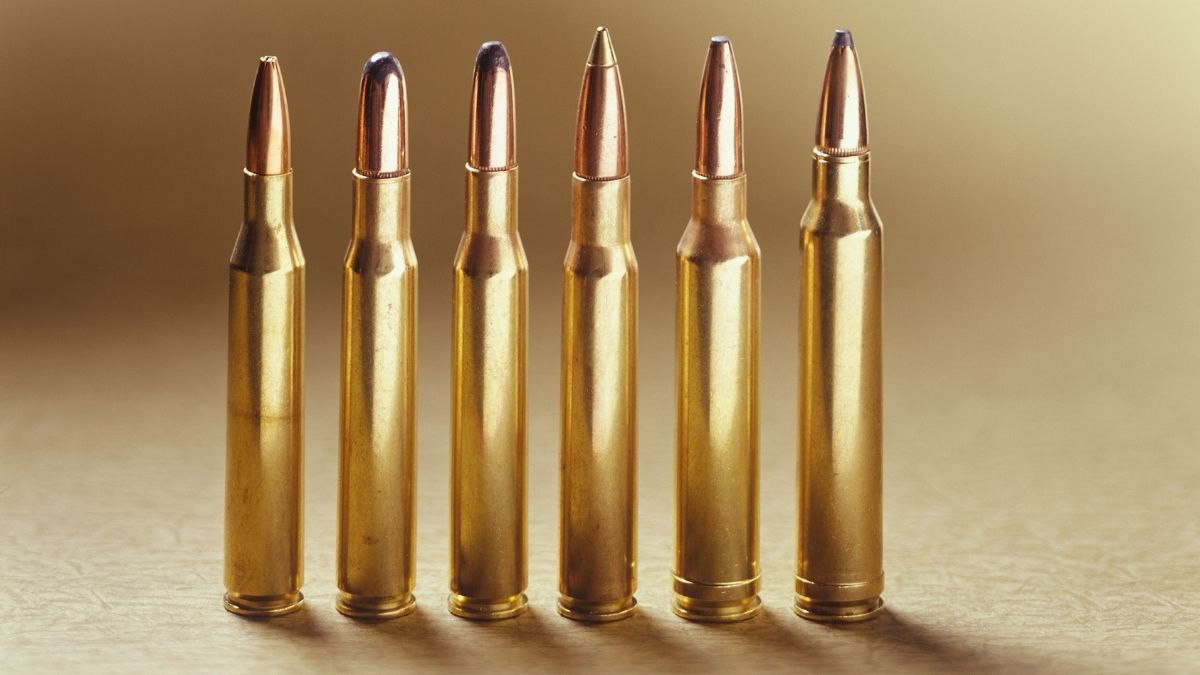 Six shiny rifle cartridges stand upright in a row on a beige surface. The lighting emphasizes their metallic sheen, conveying a serious tone.