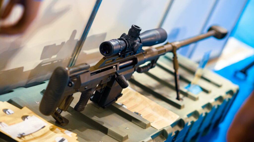 Close-up of a black sniper rifle with a scope, mounted on display. The rifle rests on a camouflage surface, with a blurred background.
