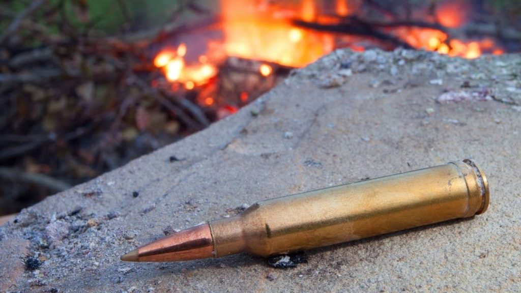A close-up of a shiny brass bullet casing resting on a rocky surface, with a blurred fire glowing in the background.
